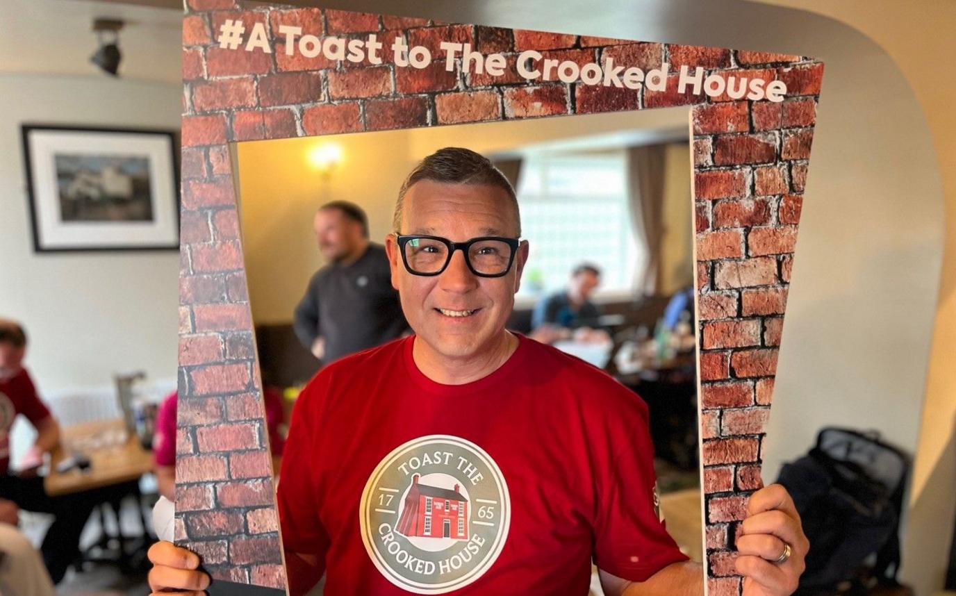 A man in a red T-shirt and glasses is looking through a piece of card that has images of bricks around the edge with a sign at the top that reads, "#A Toast to The Crooked House". There are people sitting and standing at tables behind him.