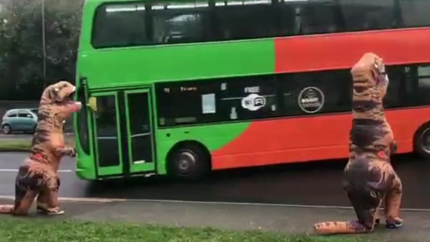 Two dinosaur costumes with a bus passing by them.