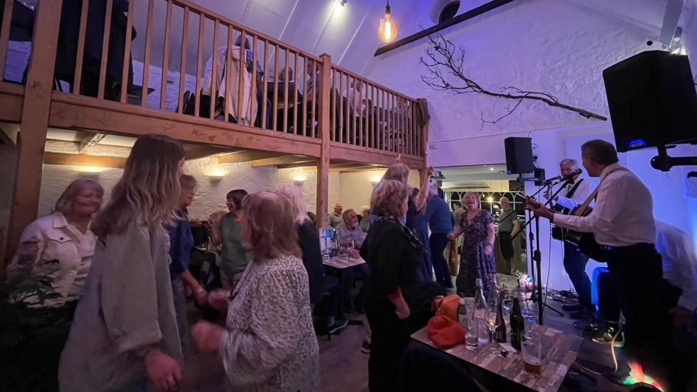 A room in a bar with a wooden mezzanine floor hosts a live music event. People are standing and sitting.