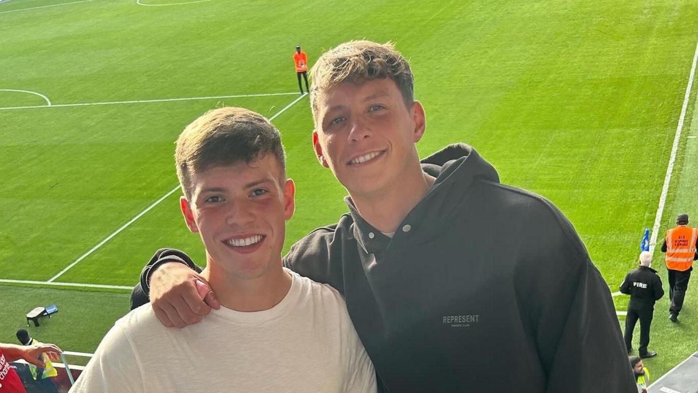 Two men stand smiling at the camera. The man on the left has on a cream T-shirt and has short brown hair. The man on the right also has short brown hair, he wears a black hoodie. Behind them is a green football pitch.