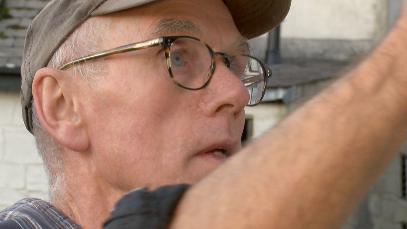 A close up of an older man with white hair, a grey cap and glasses looking to the right with his right arm raised. 