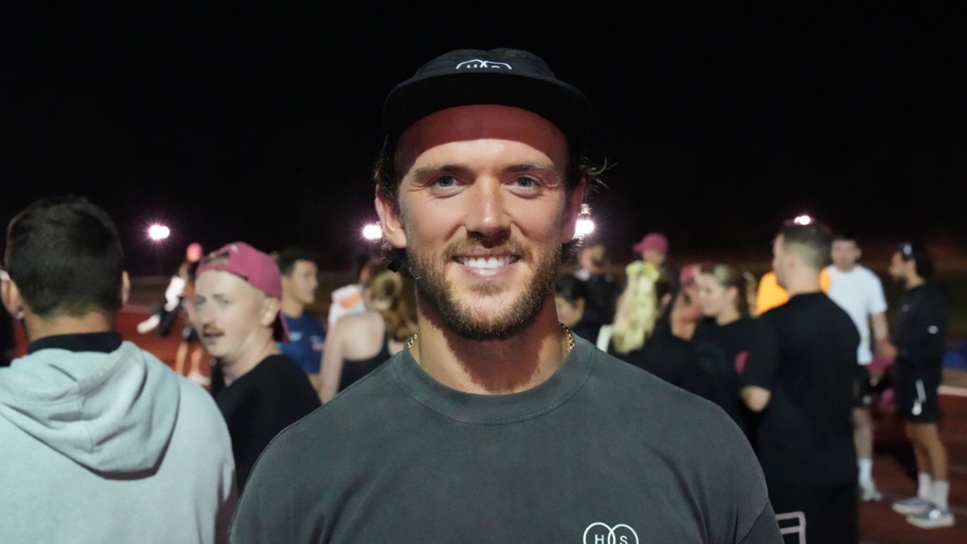 Emyr Huws is smiling into the camera. He is wearing a grey T-shirt and black baseball cap. Behind him is a crowd of other runners.