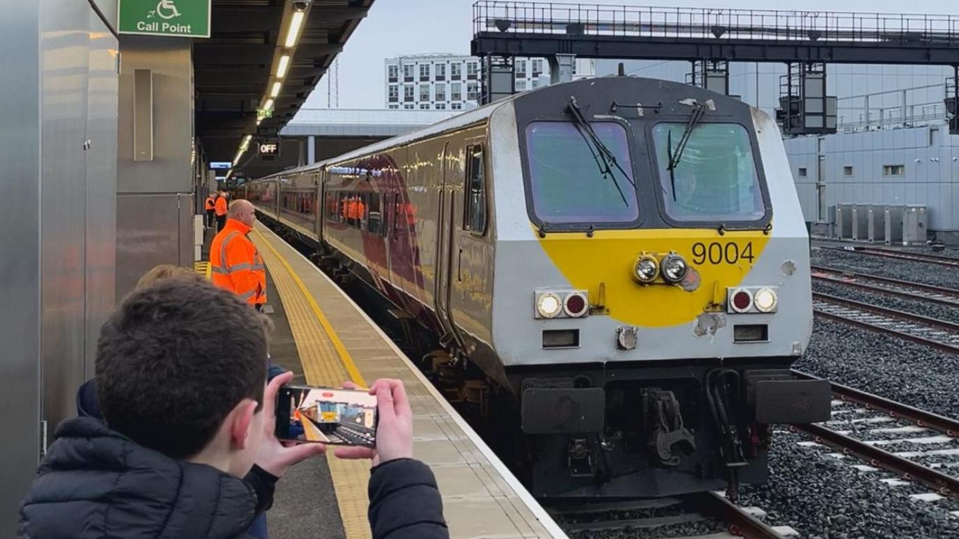 A train pulls into a station. It is yellow and grey. People stand around watching and taking pictures.