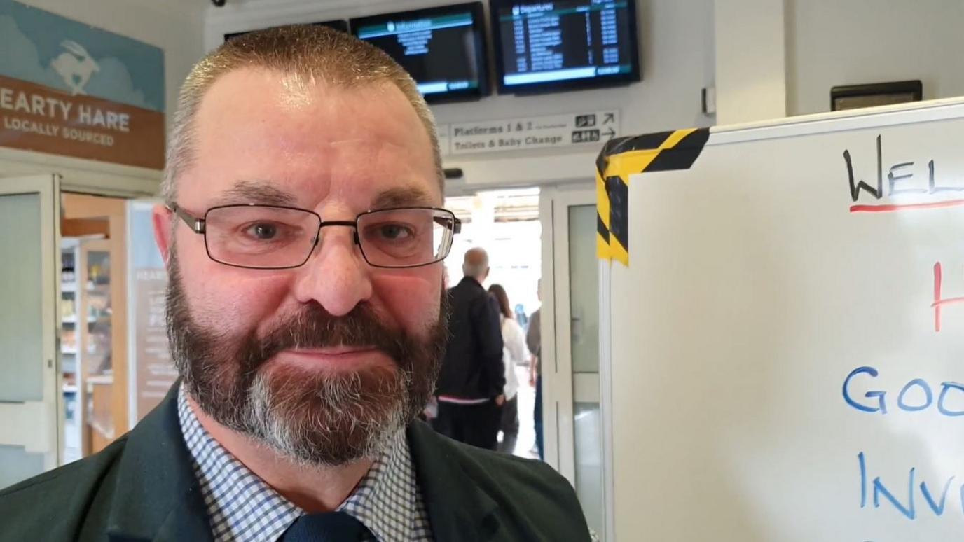 Mr Rowland wearing a gingham shirt, a dark coloured tie and blazer, and glasses. He is standing next to the whiteboard in the train station lobby