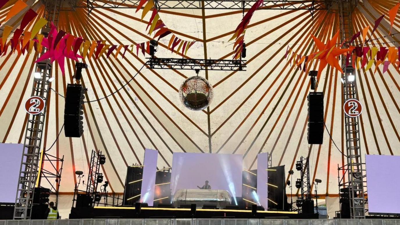 The DJ Stage inside a big tent. There are big speakers, a DJ booth in the middle with a screen behind it and a big disco ball above. There are various workers on the stage.