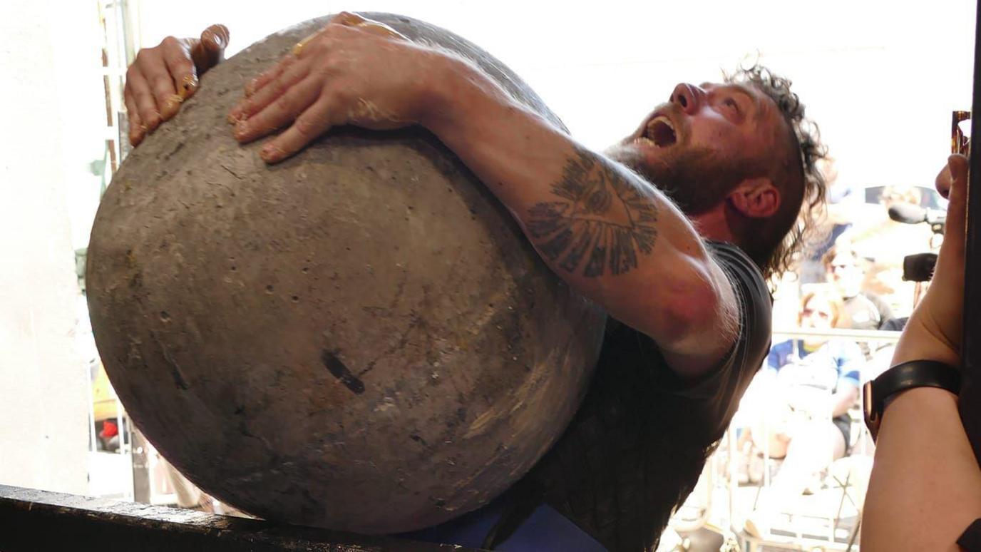 A man with a beard lifts up a huge Atlas stone and places it on a platform, looking tired with the effort of picking it up. 