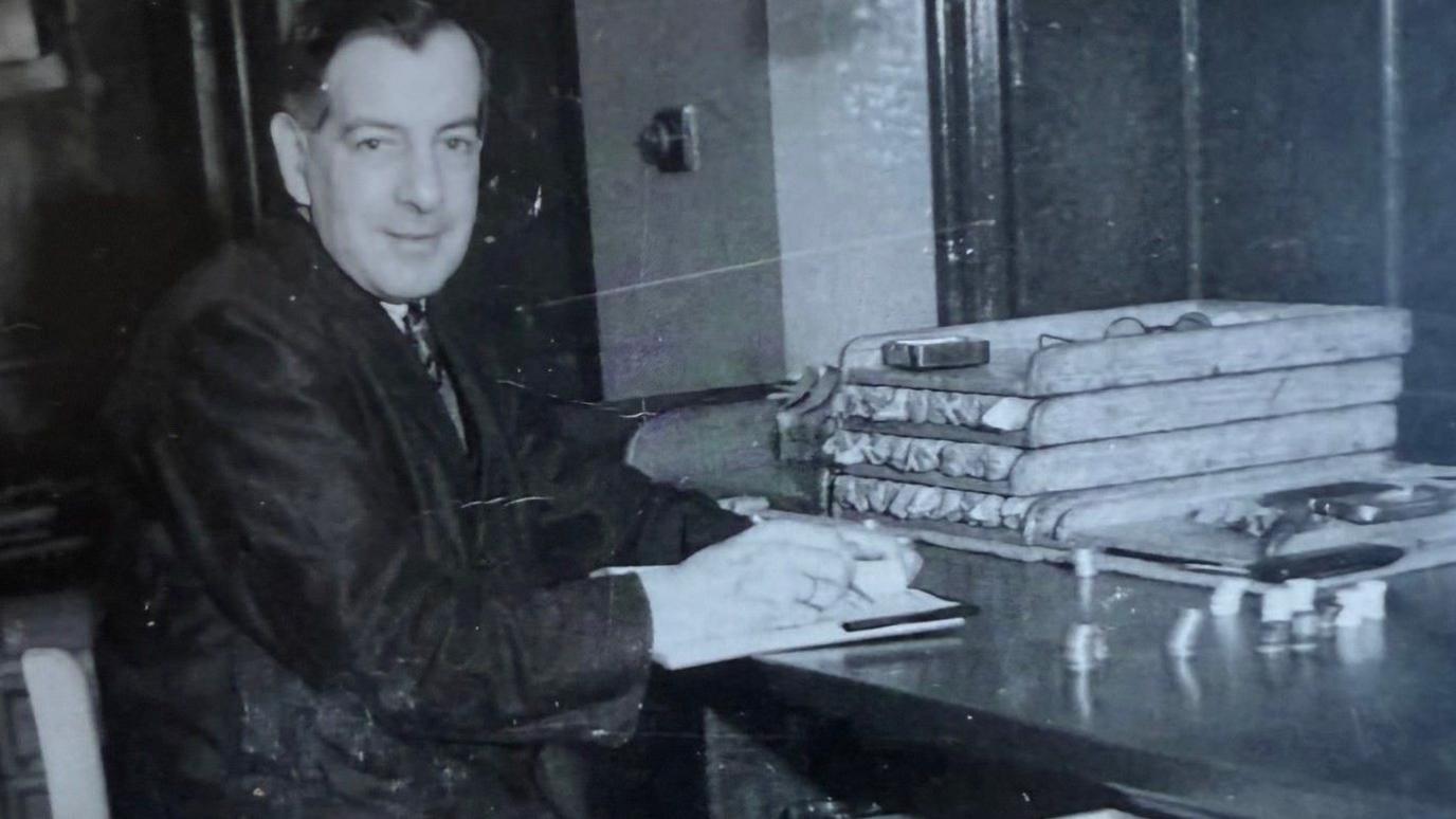 A black and white picture of a man sat at a desk wearing a suit. 