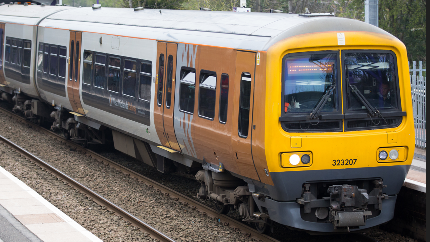 A train operatored by West Midlands Railway in Birmingham. The train has a yellow front and black and white carriages with yellow doors and shows the West Midlands Railwya logo. A platform is visible and there are overhead lines, with trees in the background.