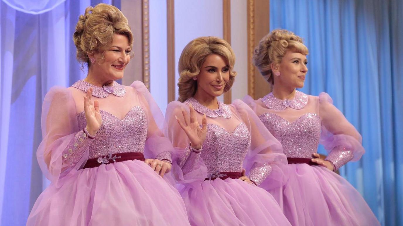 Three women wearing bouffant blonde wigs stand in a row, wearing a glittery pink chiffon long sleeve dress. Two of the women wave their hands up to the camera