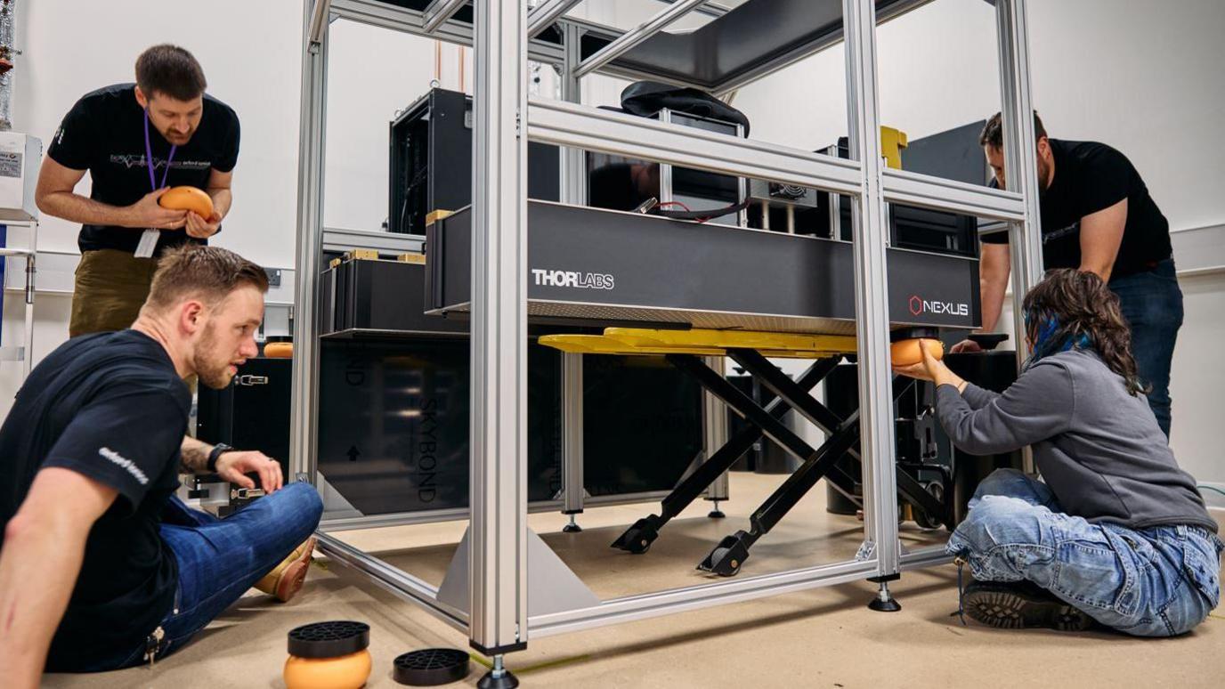 Three men and one woman of the Oxford Ionics team are working on the quantum computer installation. They are all handling different parts. None of them are looking at the camera.