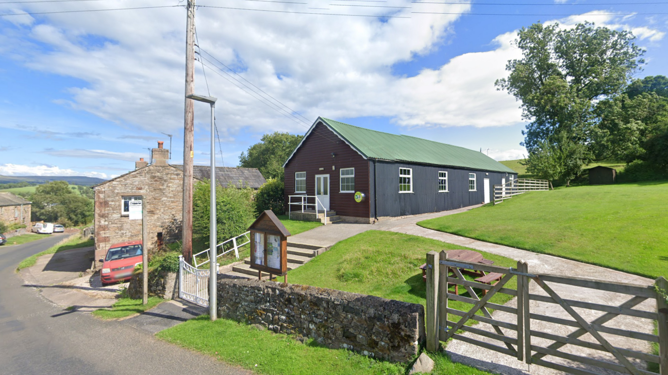 Musgrave Parish Institute is a single storey barn with a green roof and wood panelled walls. It sits on top of a grassy knoll. Down the street, you can see village houses and fields even further in the distance.