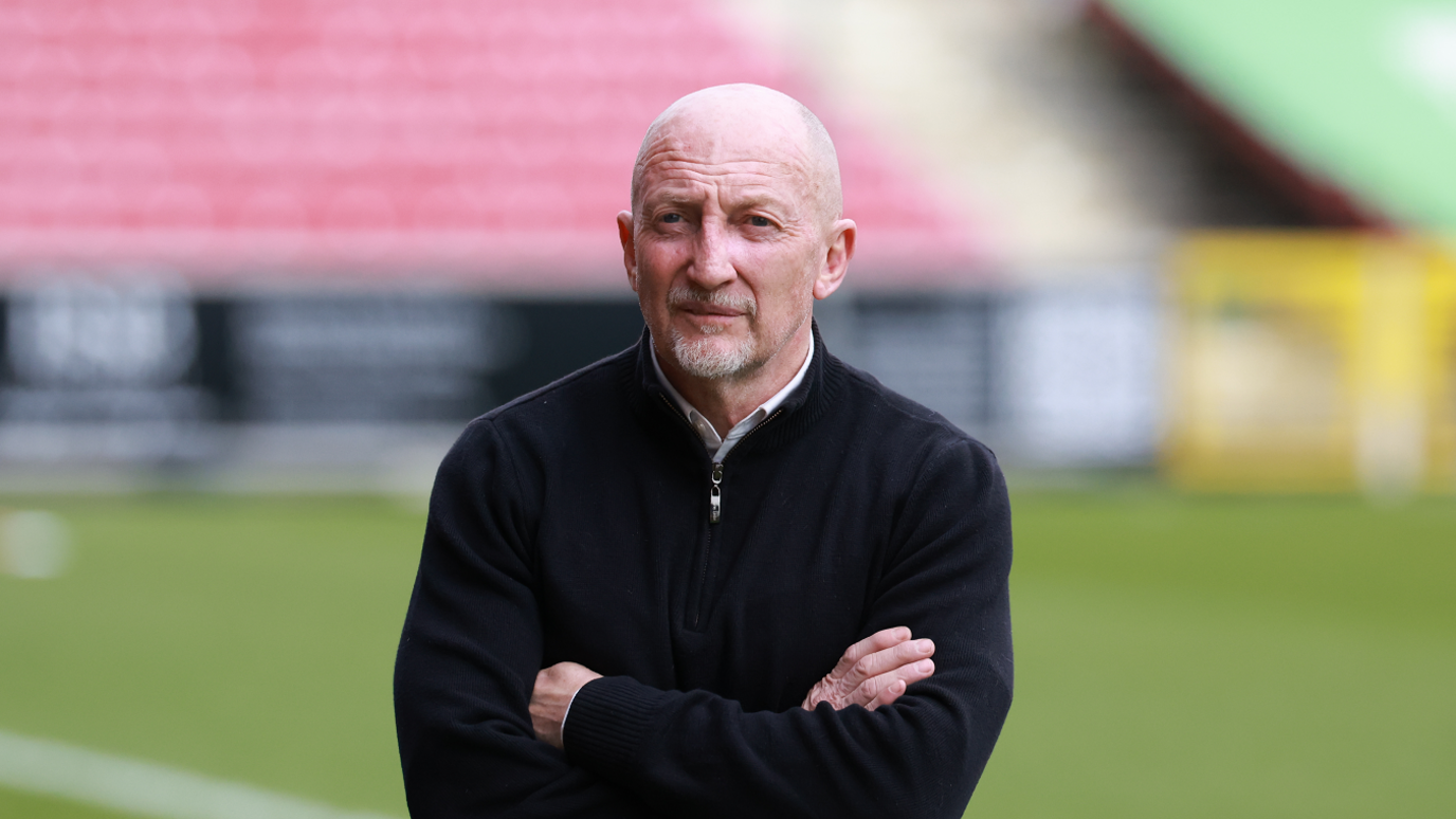 Swindon Town FC manager Ian Holloway is stood next to a football pitch with folded arms. He is wearing a black jumper over a white shirt. The background of the photo is blurred but shows red seating.