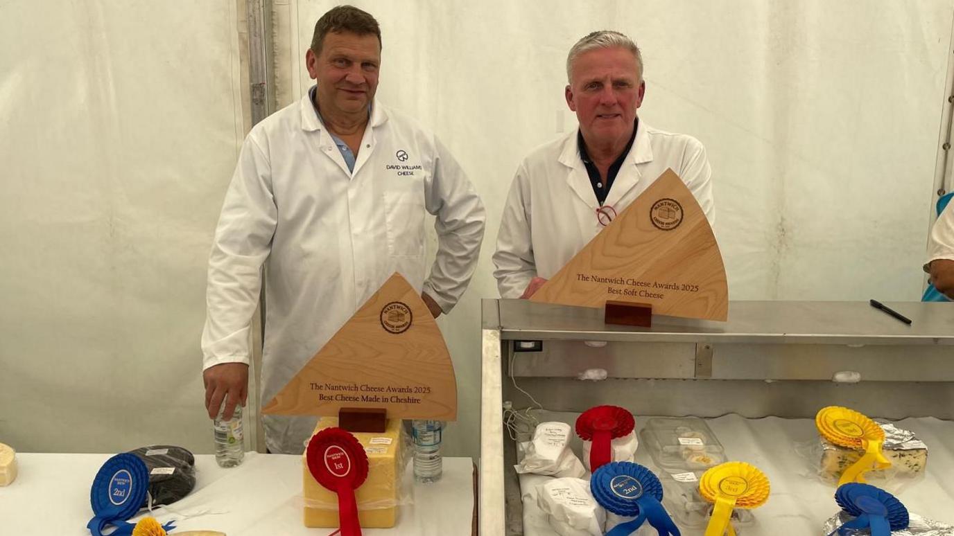 Two men in white coats stand behind a trestle table and open fridge containing cheese. Blue, red and yellow ribbons are stuck to the cheeses. There are also two wooden trophies carved into the shape of a quarter of cheese. One man has short grey hair, the other has short dark hair. They are stood inside a white marquee.