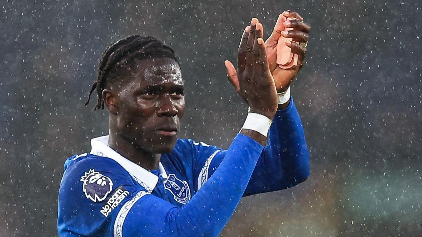 Amadou Onana, wearing Everton's blue home strip, raises his arms above his head and claps while the rain falls around him