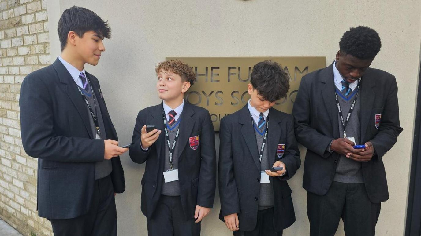 Four boys in school blazers and ties stand outside the entrance to The Fulham Boys School. They are all looking at their brick phones.