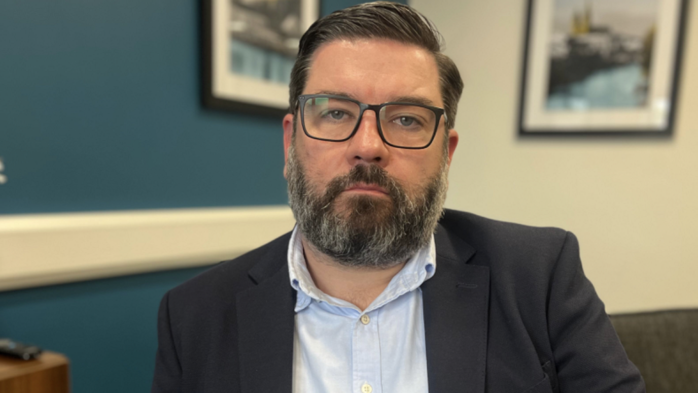 Nial McKenna, a man with dark hair and a dark beard with some grey in it. He is wearing black-rimmed glasses, a dark suit jacket and a light blue shirt.