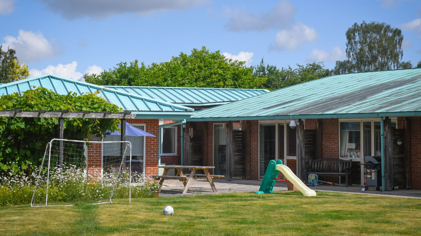 A one-storey building with green-coloured roofing on a sunny day. Trees are visible behind the building and in front is a grassy area with a football, a goal, a small slide and a picnic bench.