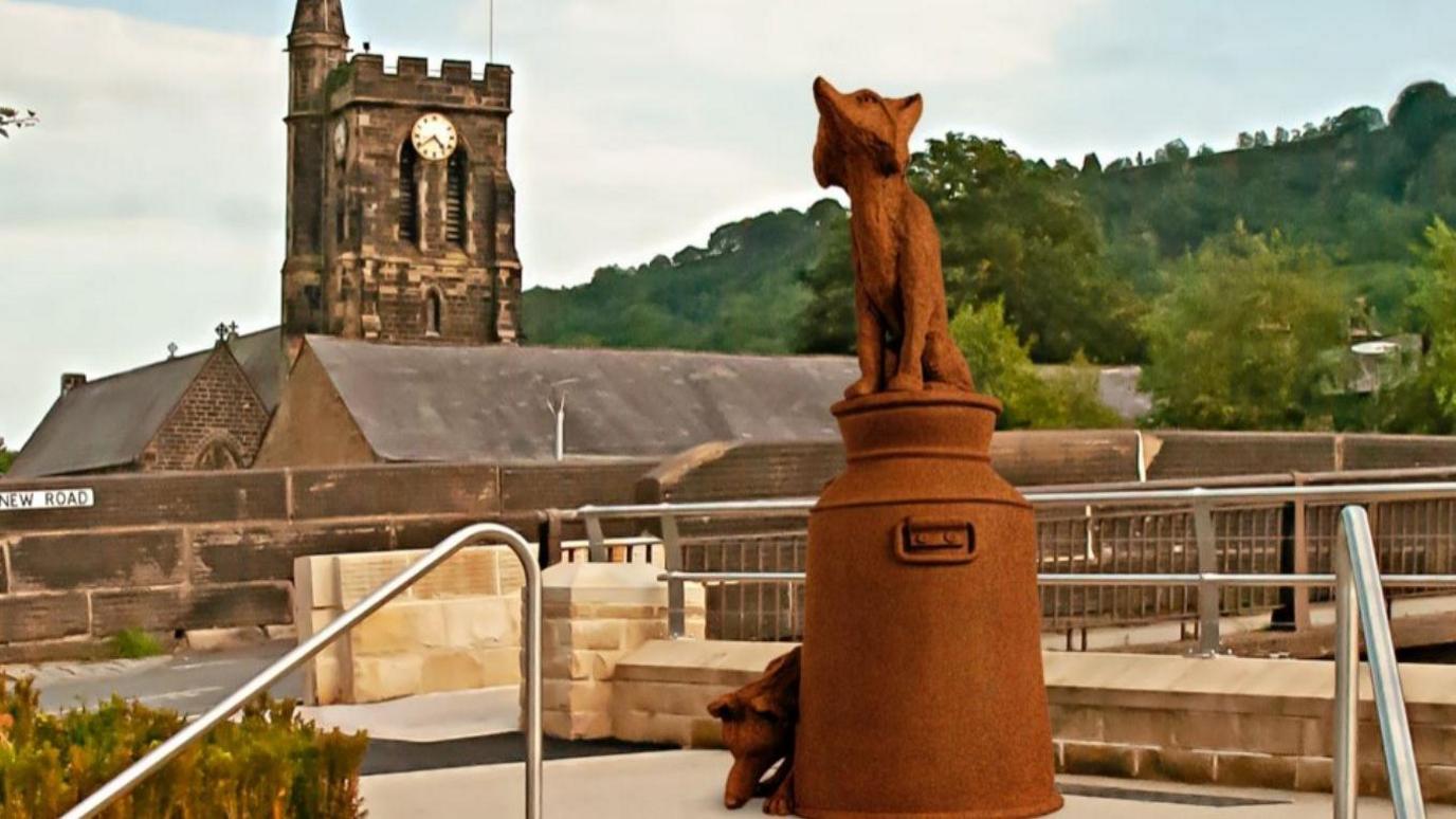 The statue is made of cast iron and depicts a fox sitting on a milk churn, with another fox next to it, sniffing the ground.