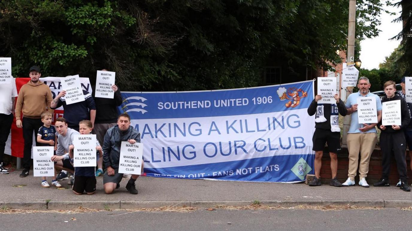 A group of men and boys holding placards urging Ron Martin to leave Southend United. They are standing either side of a large blue and white flag that accuses Martin of killing the club.