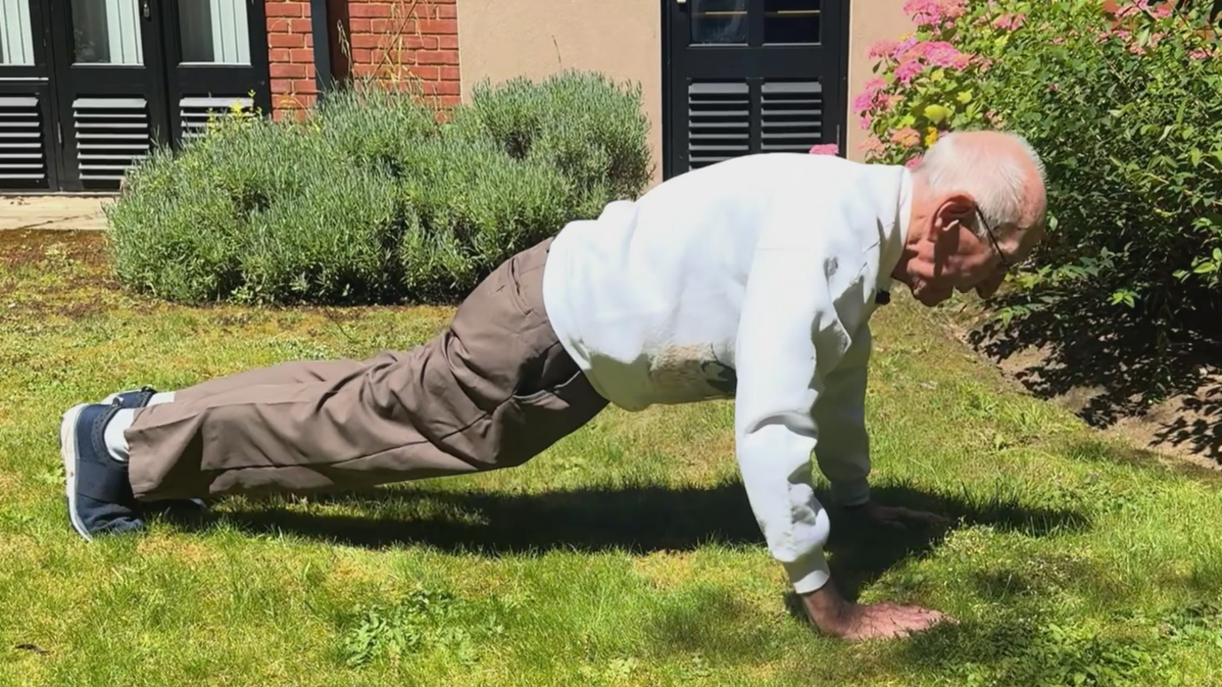 An elderly man doing a press-up on grass in a garden.