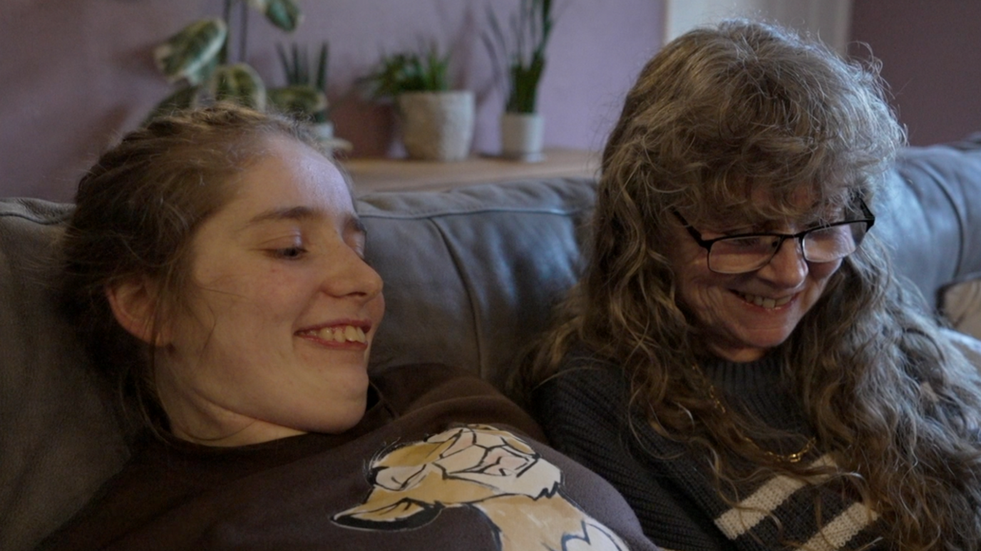 Bethany Turner and Debbie Crowson sitting on a sofa in a living room. They are smiling and looking at an electronic tablet, which is not shown in the picture.