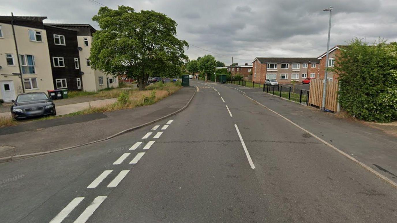 A Google street view image of a residential road. A junction is on the left and behind that is a cream and black coloured block of flats, with a car parked in the car park in front of it and a large tree. On the right are other residential buildings, fences and a bush.