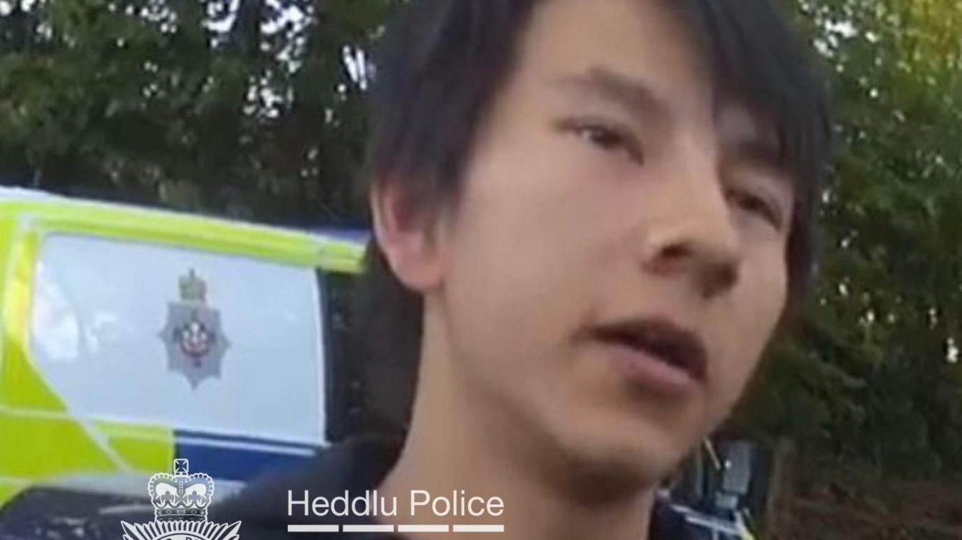 A close up shot of Jivan Dean standing in front of a police car