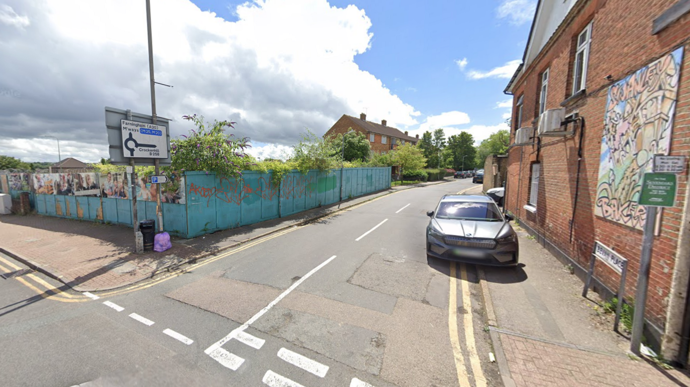 A Google Street picture of a road with a car parked on the right on double-yellow lines. There is light blue boards up blocking a site to the left of the road.