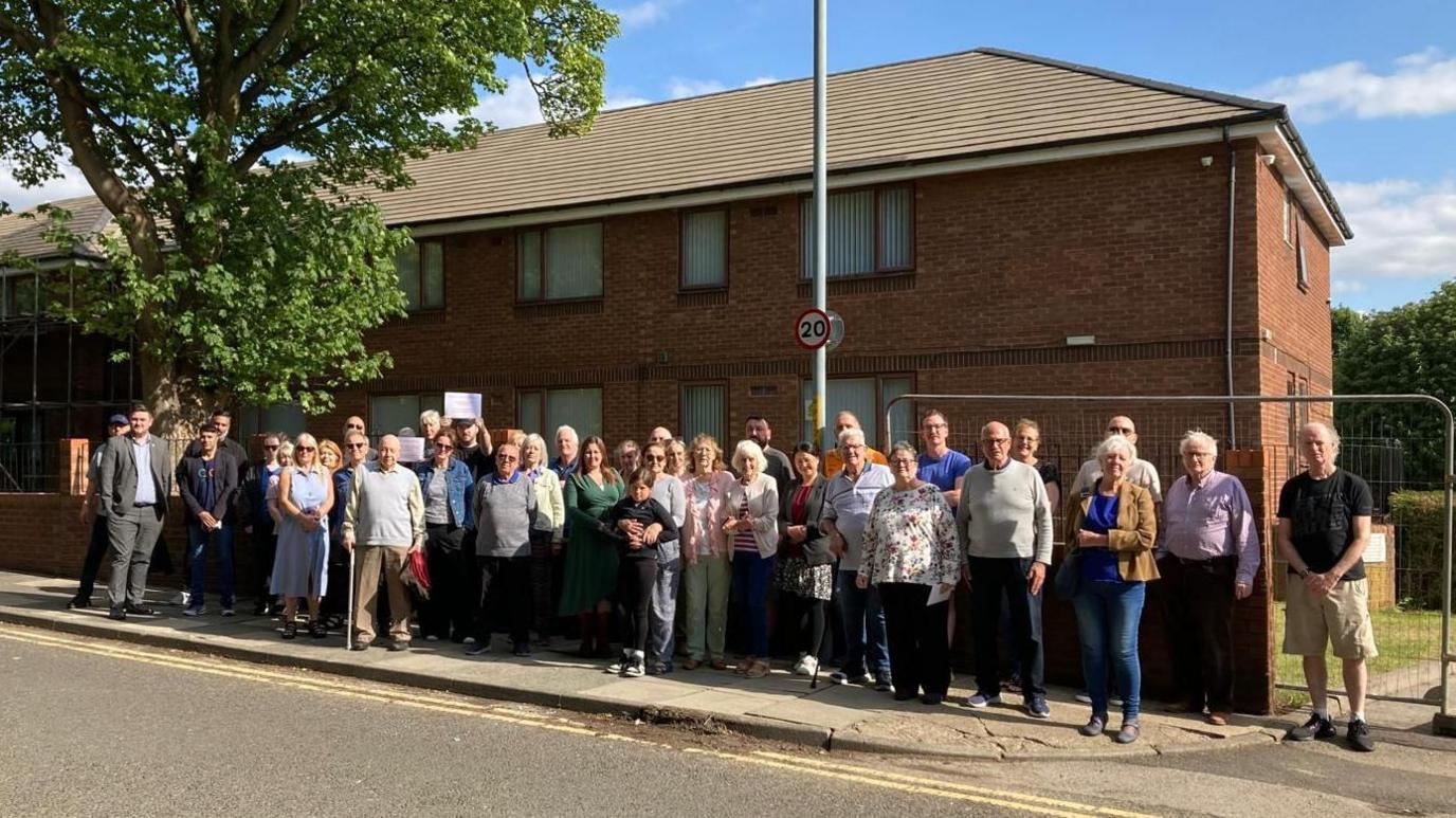 Group of 40 odd people standing outside a brick building looking at the camera. They are all dressed in various clothes. It is a sunny day.