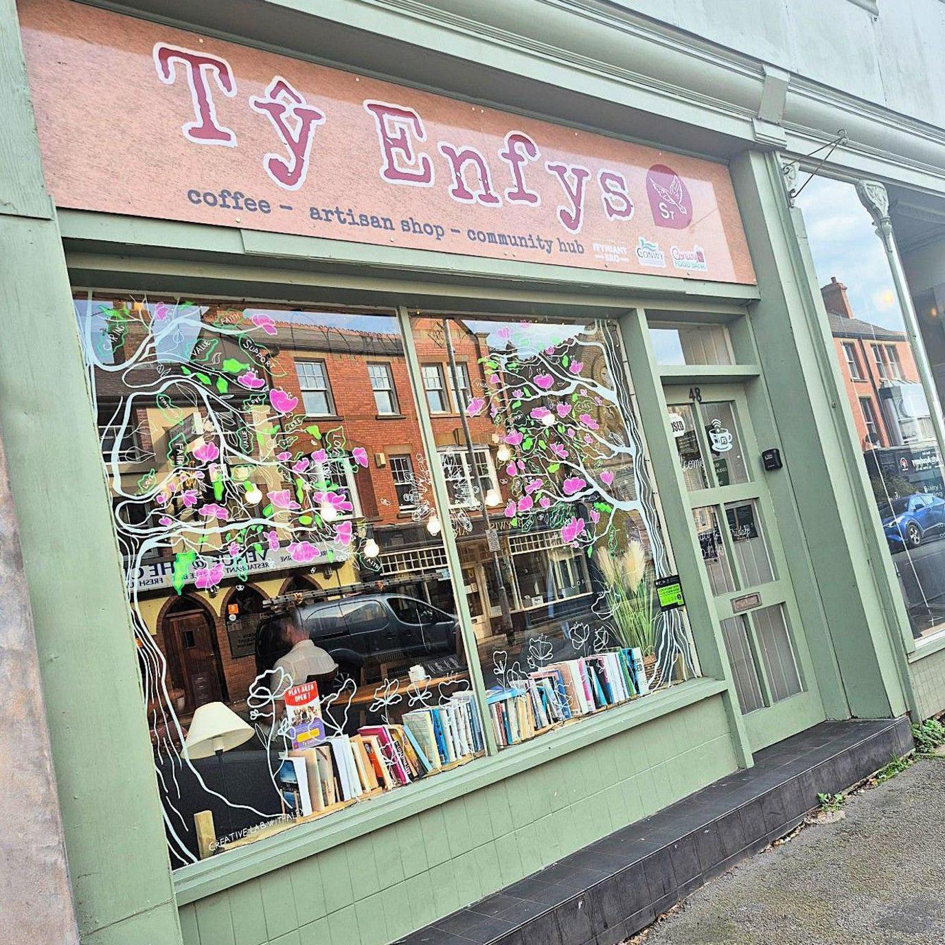 Photograph of the outside of the community hub, labelled Ty Enfys. The building is sage green and there are pink flowers painted on the windows.