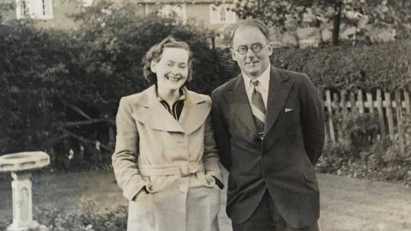 A black and white photo of a Elva and Anthony Pratt on a lawn. There is a bird bath behind them to the left and a hedge beyond that. Anthony Pratt is wearing dark-rimmed circular glasses and a suit. His wife (to the left is in a light coloured coat