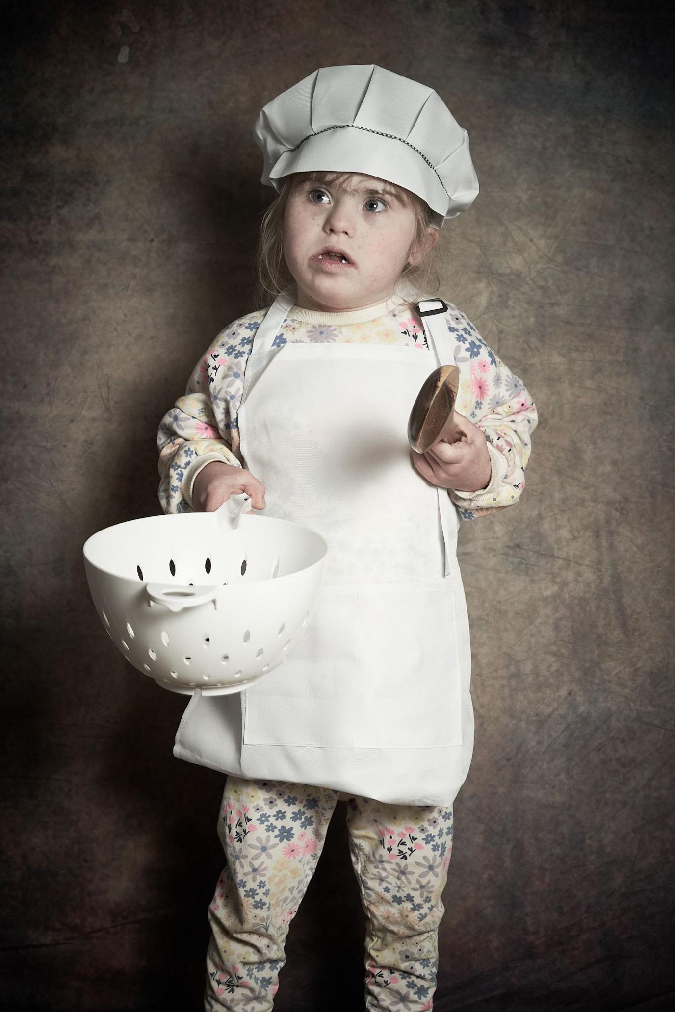 Lacy-Mai, a young girl with brown hair, is wearing a white chef's hat and white apron over her floral jumper and trousers. She is holding a large wooden spoon in one hand and a white colander in the other.