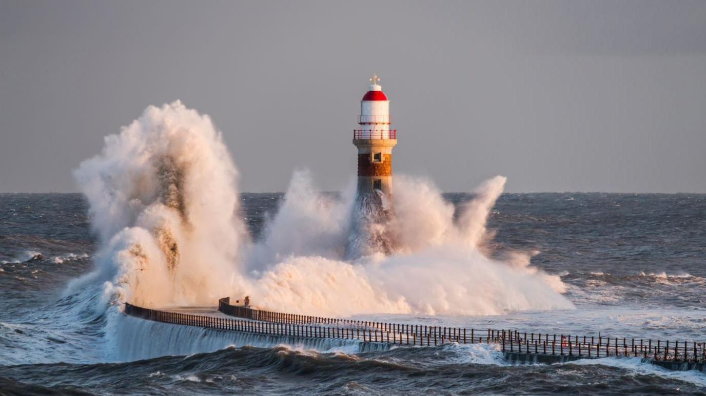 Roker Pier. Large waves are crashing into the pier and it's lighthouse. The pier curves to the right and has railings along each side. The lighthouse has a white and red dome.