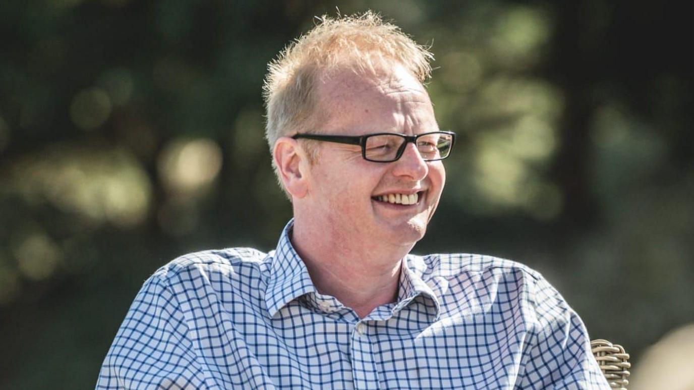 Leo Morris, wearing a checked shirt and glasses, sits in a chair and smiles.