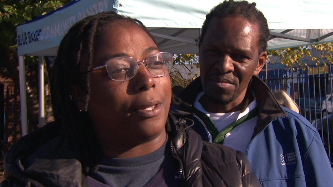 Winnie, wearing a black bubble coat and glasses, speaks to the camera with a male companion in a blue coat looking on. 