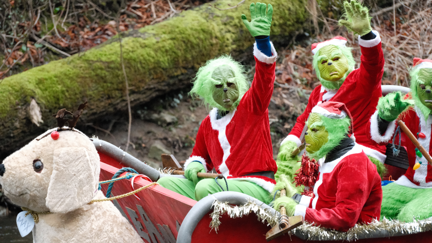 People dressed in green Grinch masks and red santa outfits waving towards the camera from the top of a raft