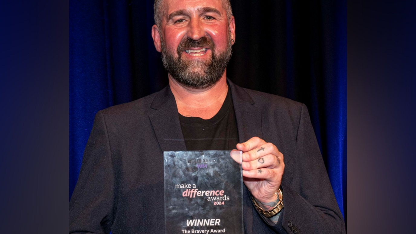 Steve Whitehouse is wearing a black suit with a T-shirt underneath and is smiling holding his award in the photo. Steve has dark hair and a beard and is wearing a gold-coloured watch on his left hand and his fingers are tattooed.