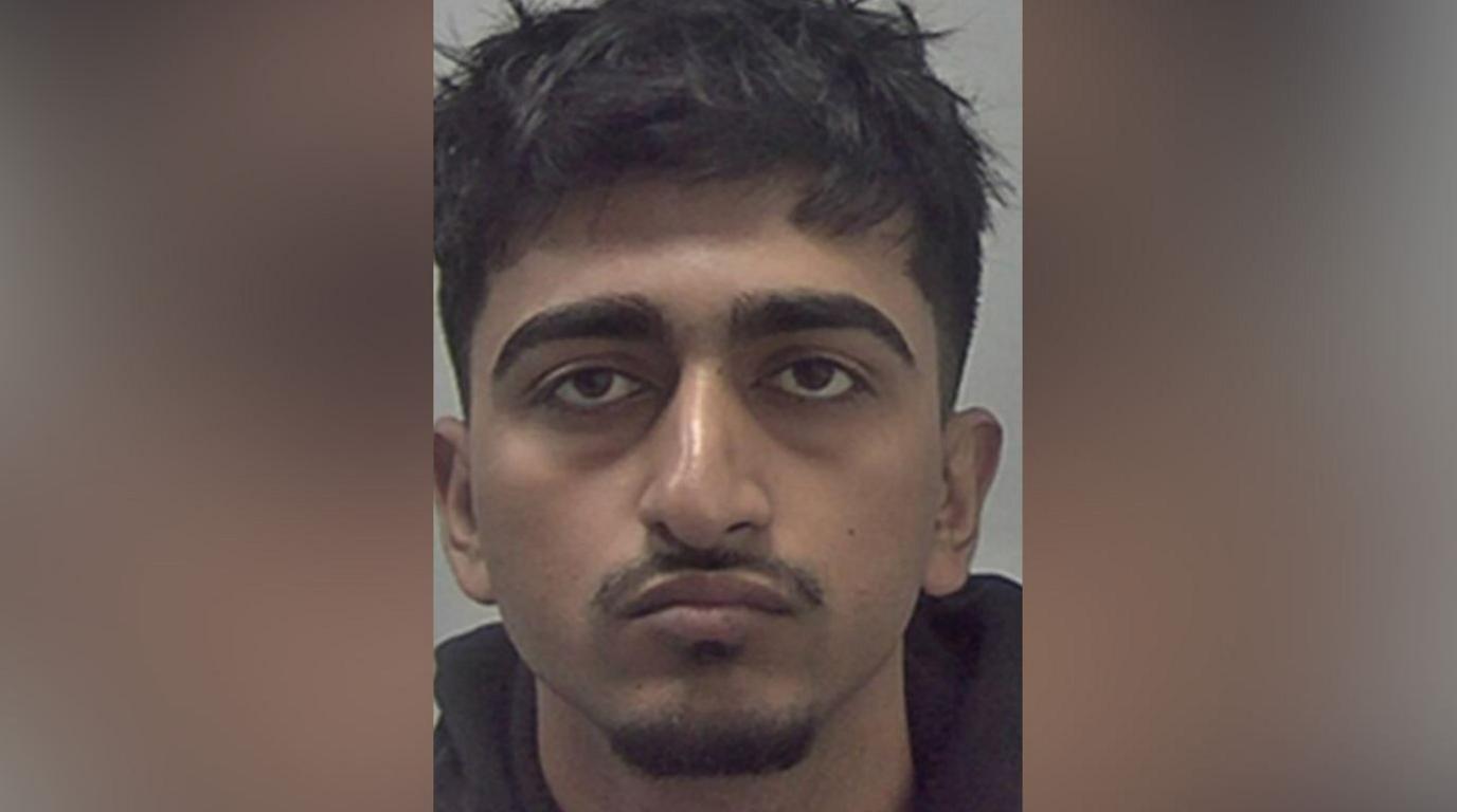 A mugshot of a man with messy black hair and a moustache and goatee beard. He is wearing a black top.