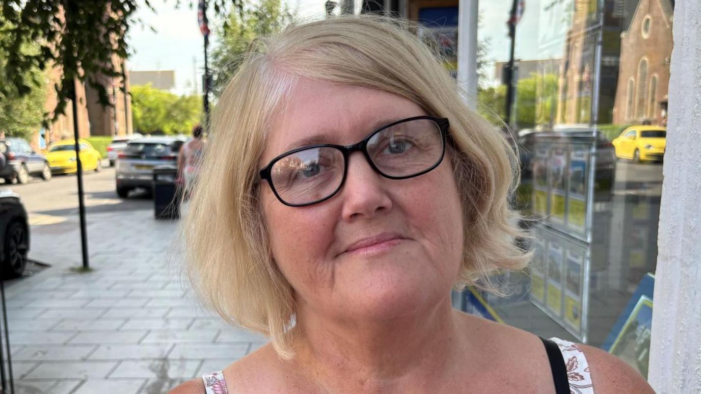 Denise Webb has blonde hair and black glasses. She is wearing a vest top on a paved street.