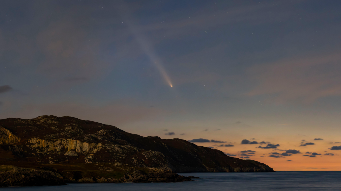 Sunset overlooking cliffs with reddish skies on the horizon as it meets the sea, and in the centre of the image is a streak of light from a comet 