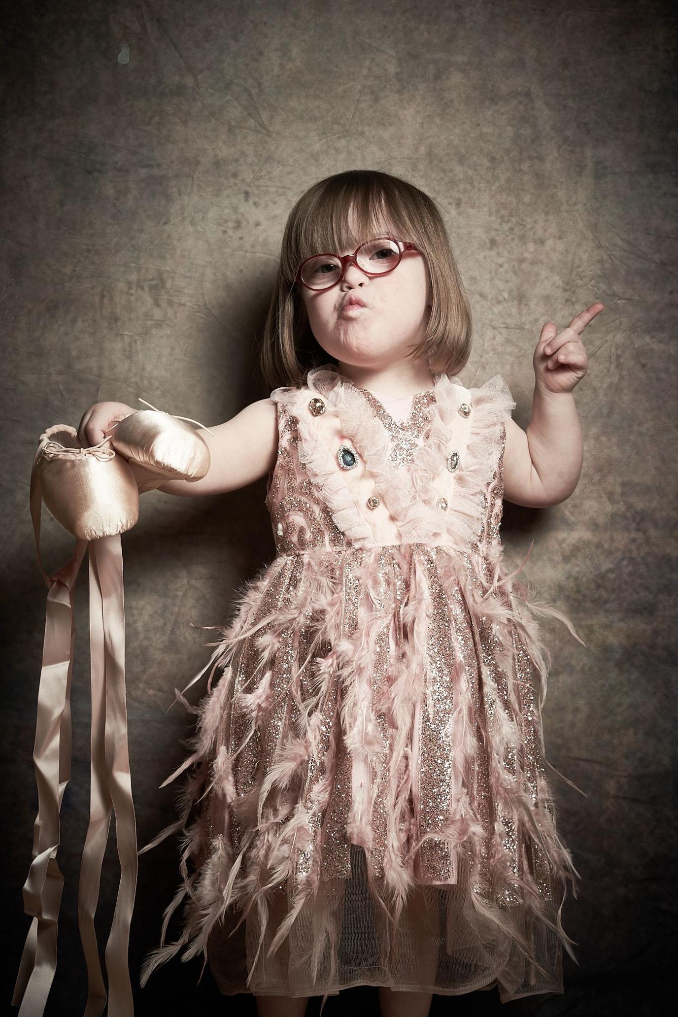 Freya, a young girl with short brown hair and glasses holds out ballet shoes. She is pointing with her other hand and has a sassy pout on her face.