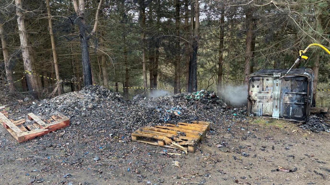 A large pile of scorched lithium-ion batteries that had been discarded near woodland. Two wooden pallets are also amongst the waste pile and a metal crate on its side is on the right of the frame, with a thin yellow tube misting down the scene.