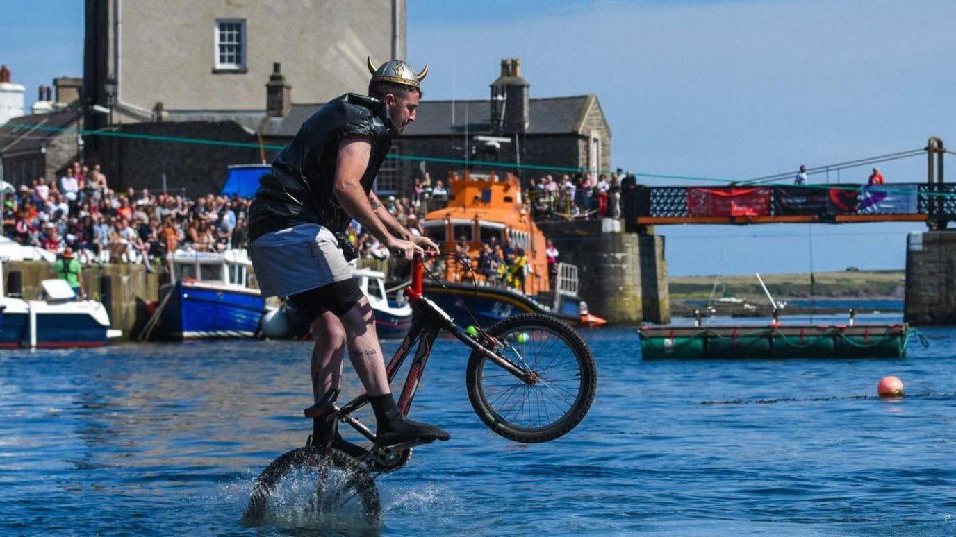 A man jumps into the sea riding a bike, dressed in a bin bag with a silver and gold helmet with horns. He is mid-flight and his back wheel splashes into the water. A busy crowd watch on from the harbourside. An orange RNLI boat can also be seen in the background.