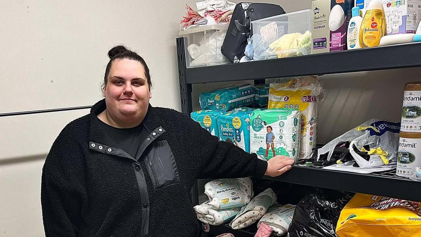 Megan Sinden, whose black hair is in a ponytail, is standing in front of shelves stacked with baby products including nappies and formula.