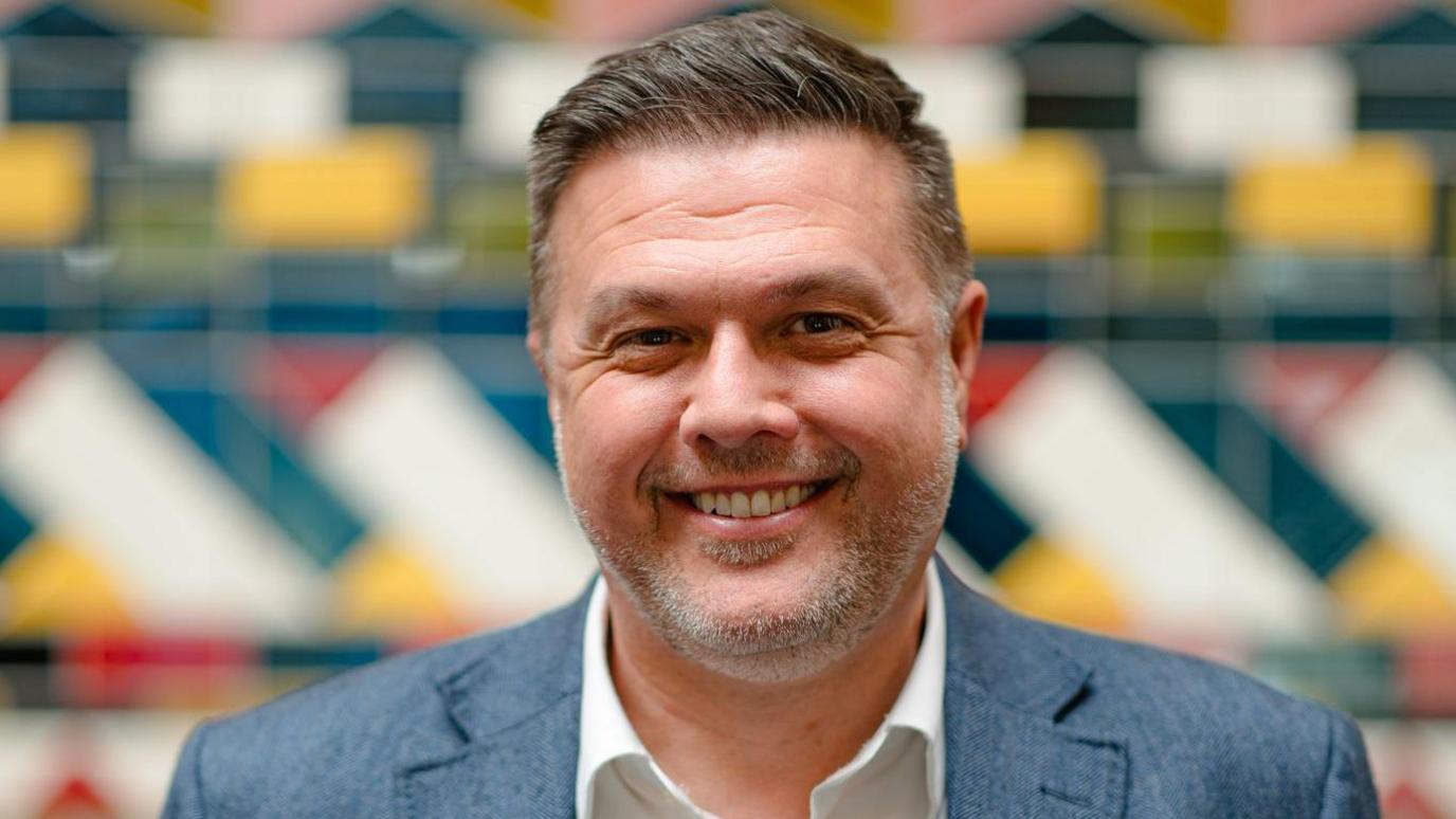 Simon smiles at the camera. He has short brown/grey hair and light grey stubble, and wears a white shirt and grey suit. He is standing in front of a blurred, multi-coloured background. 