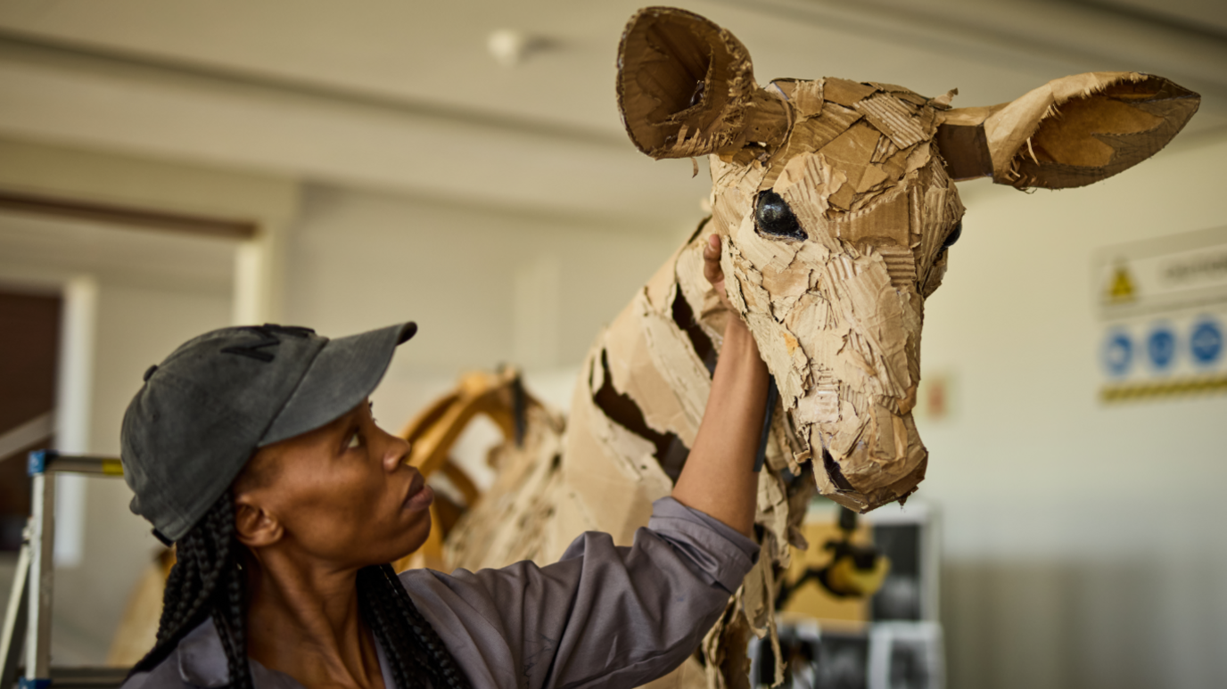 The head section of a giraffe puppet is shown close-up, with one of the operators resting her hand on its necks