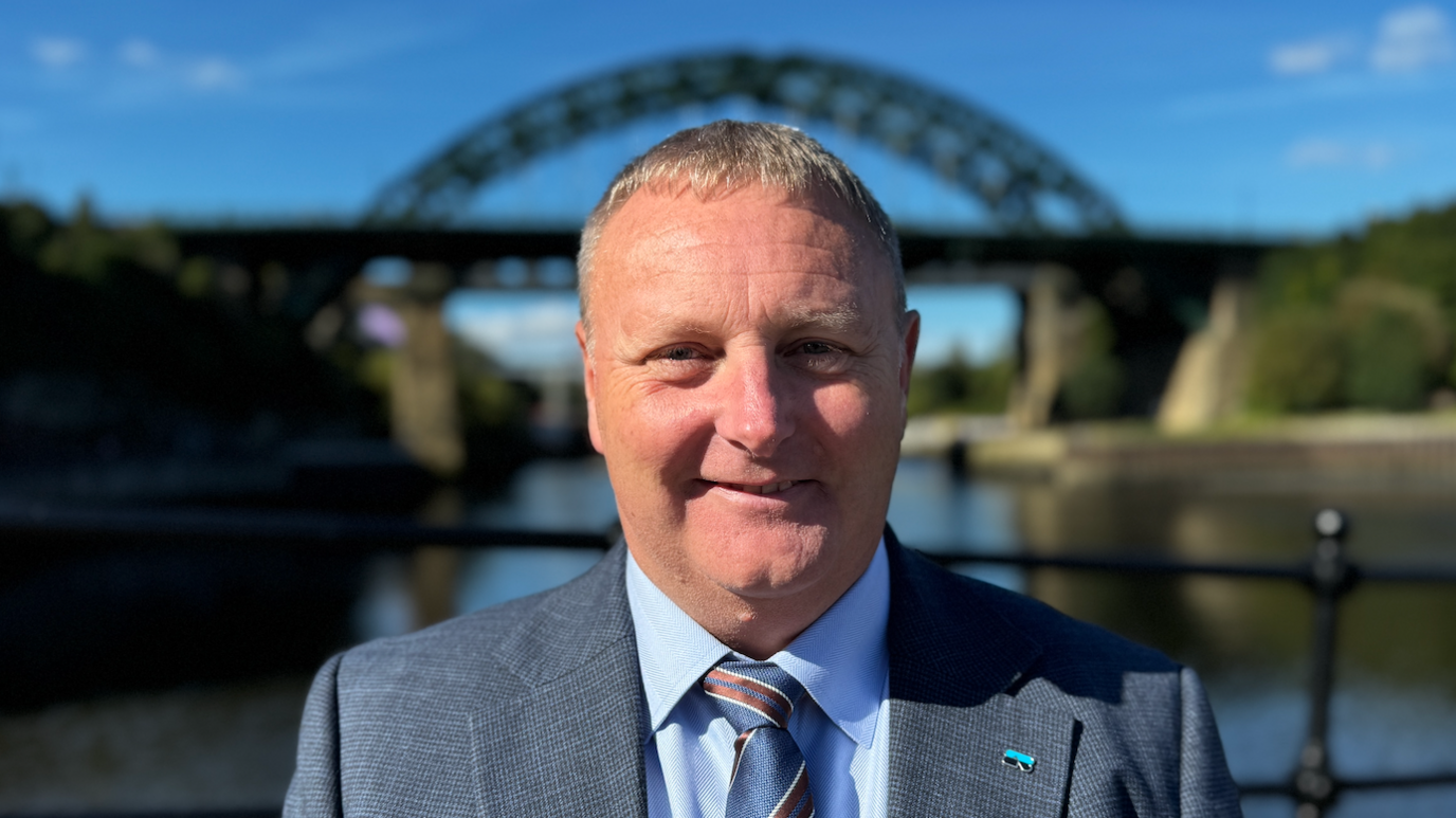 Ian Cussons standing with the a bridge in the background which his blurred. He is smiling and wearing a blue shirt, tie and jacket.