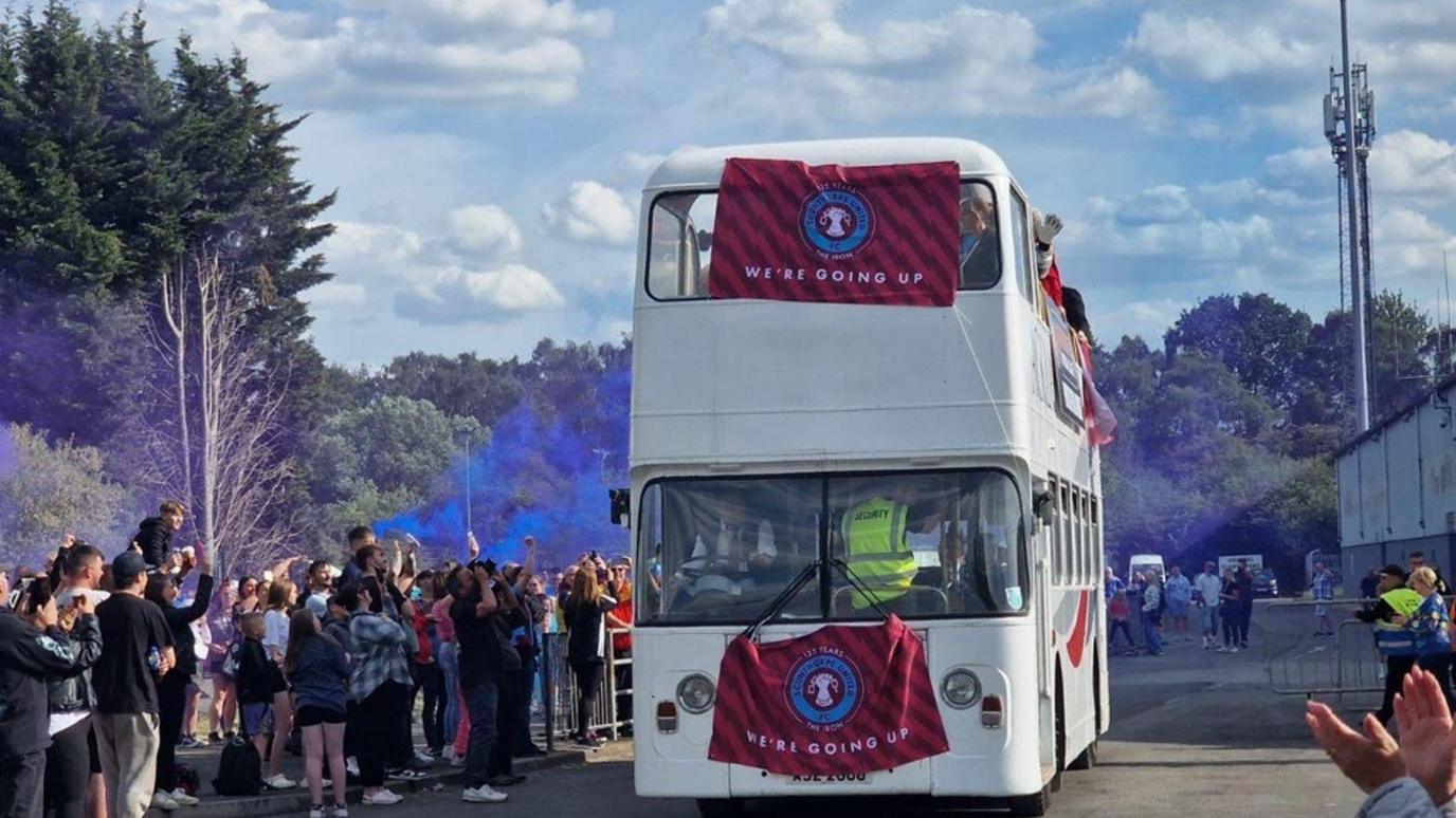 A white double decker bus makes its way past a crowd of people with Scunthorpe United flags stating 'we're going up' draped at the front of it. Blue powder can be seen going through the air.