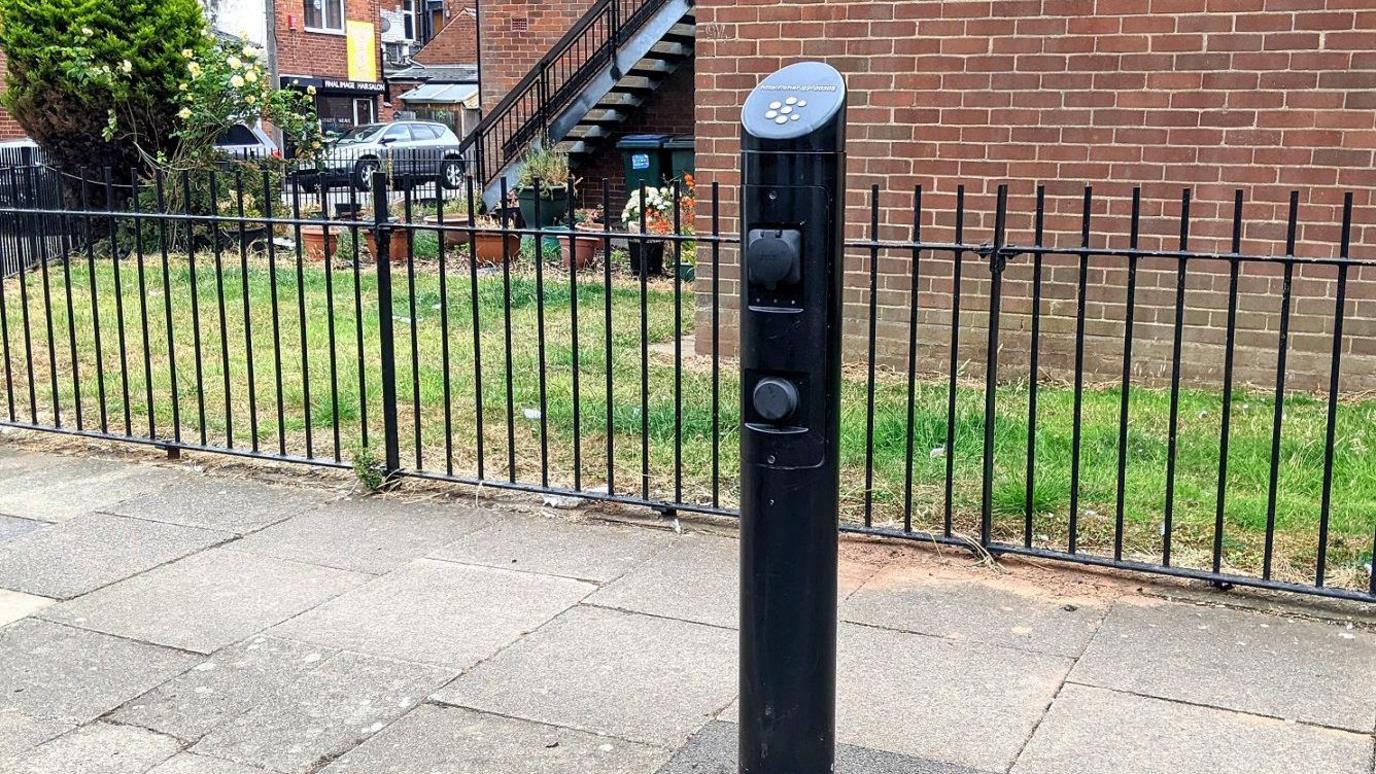 An electric car charging point in Coventry has a black post in the pavement. It is in a residential area and there is a fence behind the pavement with a lawn and steps leading up to a building. Cars are parked in a road in nearby.