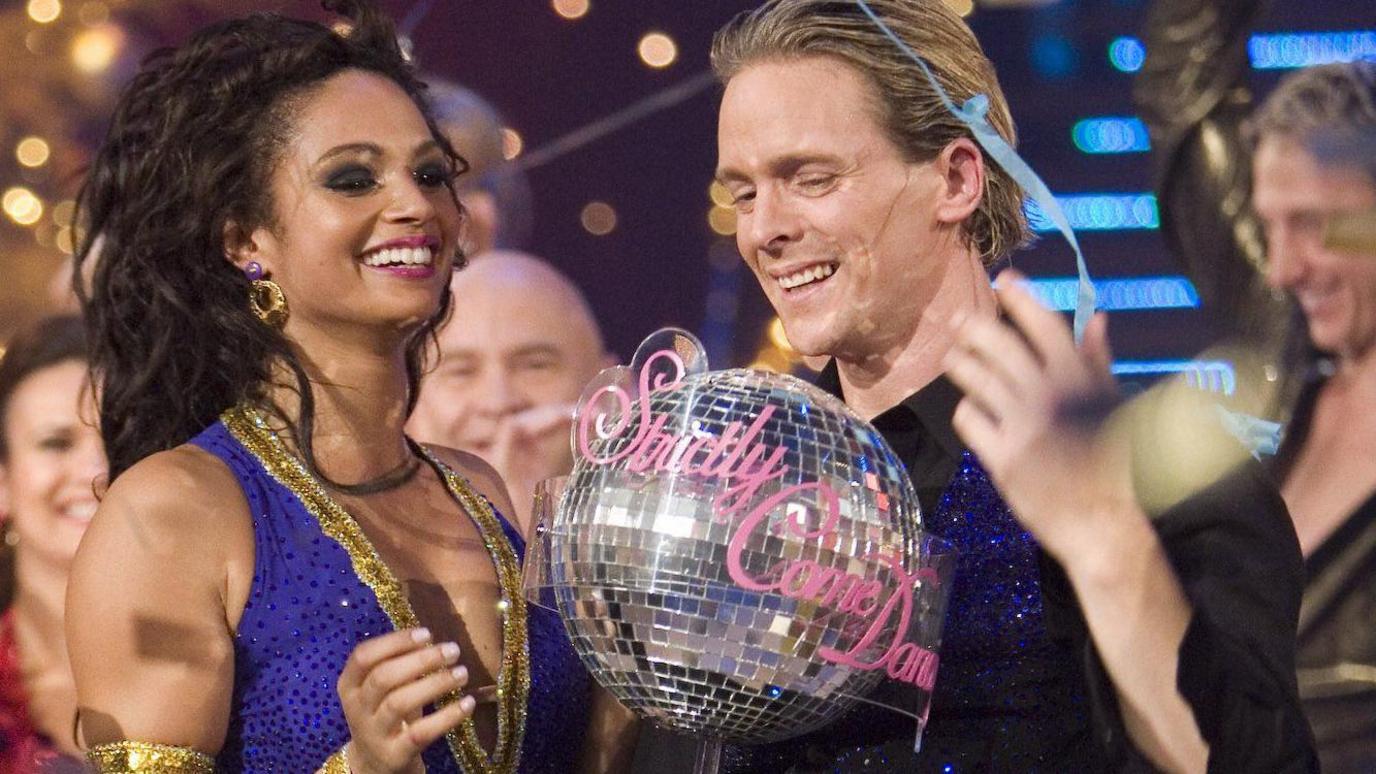 Alesha Dixon with long curly black hair and wearing a midnight blue and gold sleeveless costume holding the glitterball trophy with Matthew Cutler, who had fair hair swept back and is wearing a black shirt and midnight bue sparkly waistcoat.